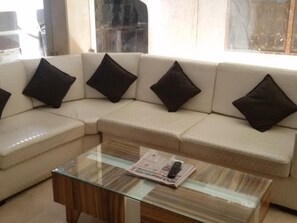 A living room with a beige sectional sofa, black cushions, and a glass-top coffee table with a newspaper and remote control. - Oly Saffron Near New Delhi Railway st. (New Delhi)