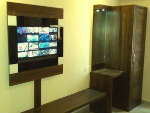 Una habitación con una televisión instalada en la pared mostrando varios canales, un armario de madera y un banco.