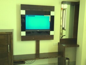 A television mounted on a wall with a wooden frame, a mirror above it, and a power outlet below. - Oly Saffron Near New Delhi Railway st. (New Delhi)