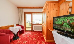 Ein Hotelzimmer mit Fernseher, roten Polsterstühlen, einem kleinen Tisch und Blick ins Freie durch eine Schiebetür.