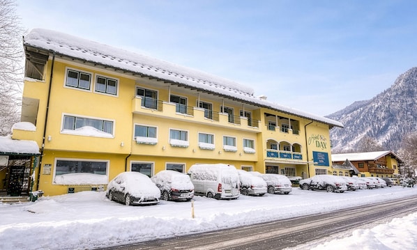 A multi-story hotel with a yellow facade, several balconies, and a parking area with snow-covered cars. - Hotel Dax by Alpeffect Hotels (Lofer)