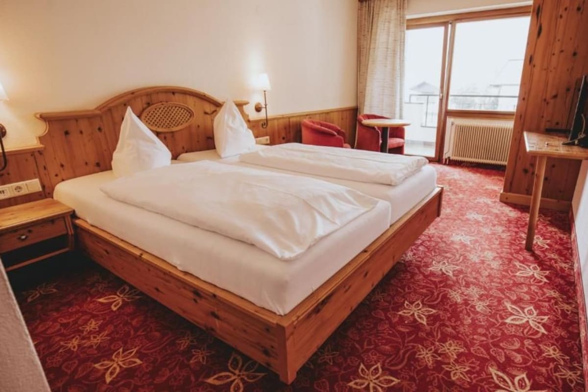 A hotel room with a wooden bed, a red carpet, a small table, and a window with a view.