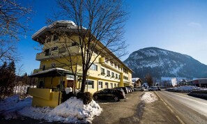 A multi-story hotel with a snow-covered ground, parked cars, and a mountain in the background. - Hotel Dax by Alpeffect Hotels (Lofer)