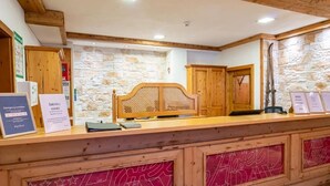 A reception desk with a wooden headboard, stone wall backdrop, and emergency contact signs. - Hotel Dax by Alpeffect Hotels (Lofer)