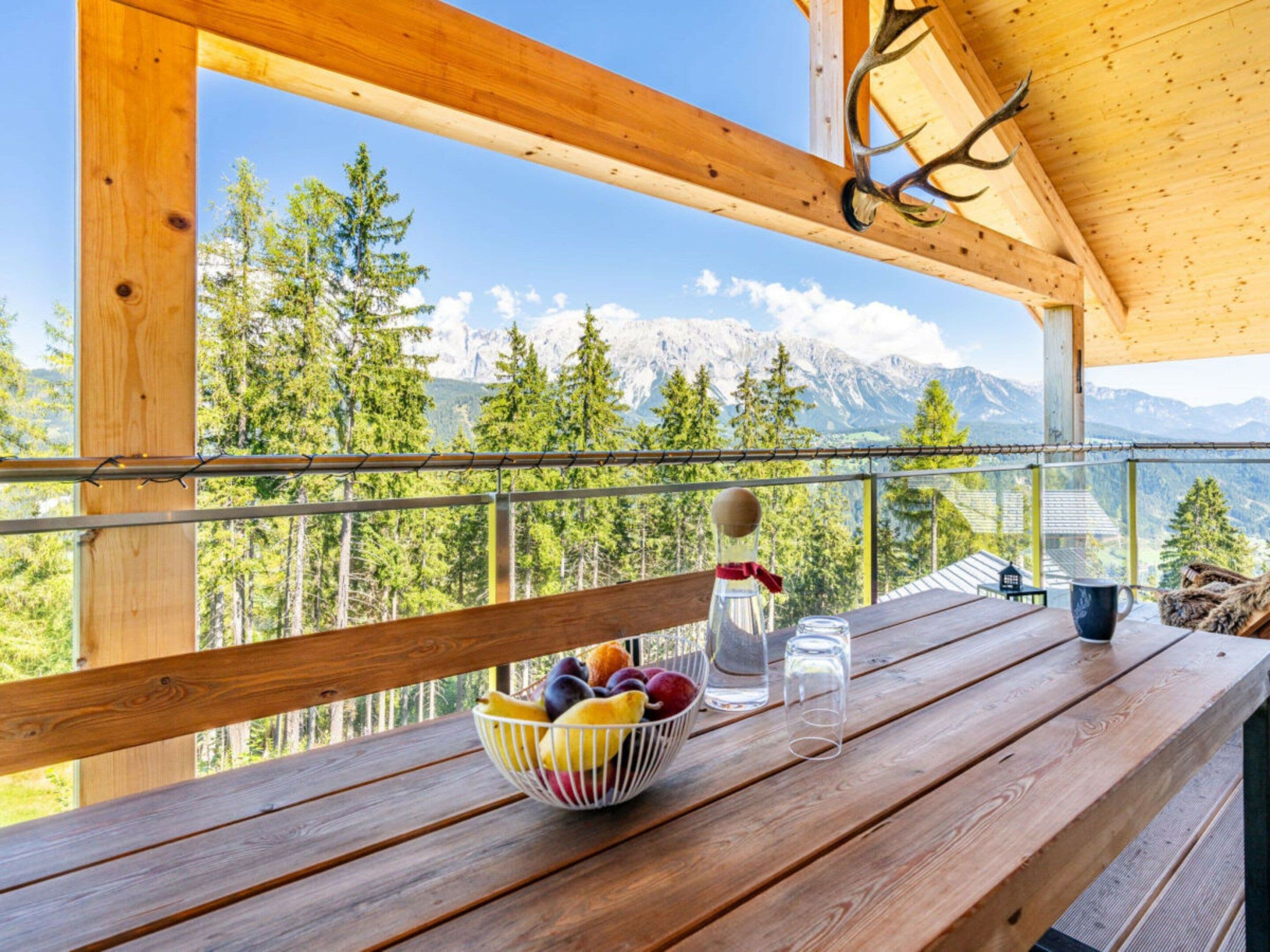 Une terrasse en bois avec une table, un saladier rempli de fruits, une bouteille et des verres. Une branche de cerf est suspendue au plafond.