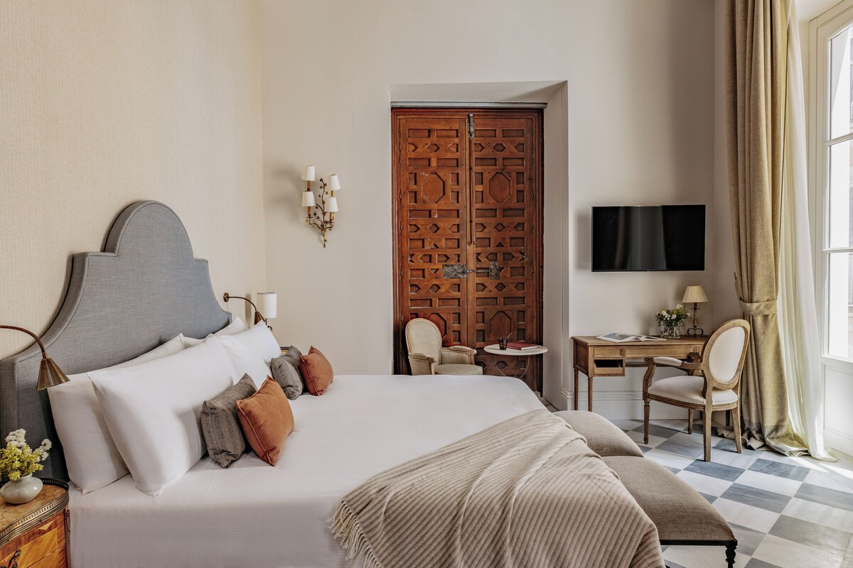 A hotel room with a large bed, a desk, a chair, a television, and a wooden door.