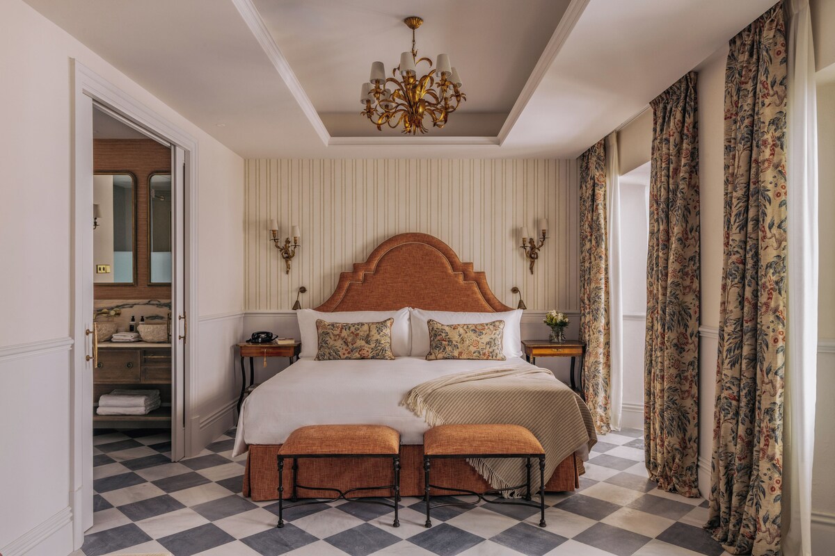 A hotel bedroom with a checkered floor, a large bed with a wooden headboard, two bedside tables, a chandelier, and a view of a bathroom through an open door.