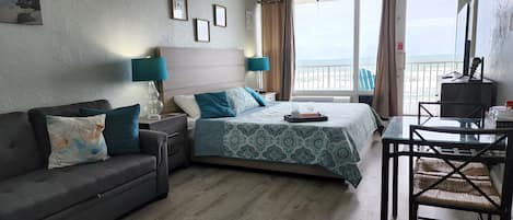 A hotel room with a bed, a sofa, a desk, and a view of the sea.