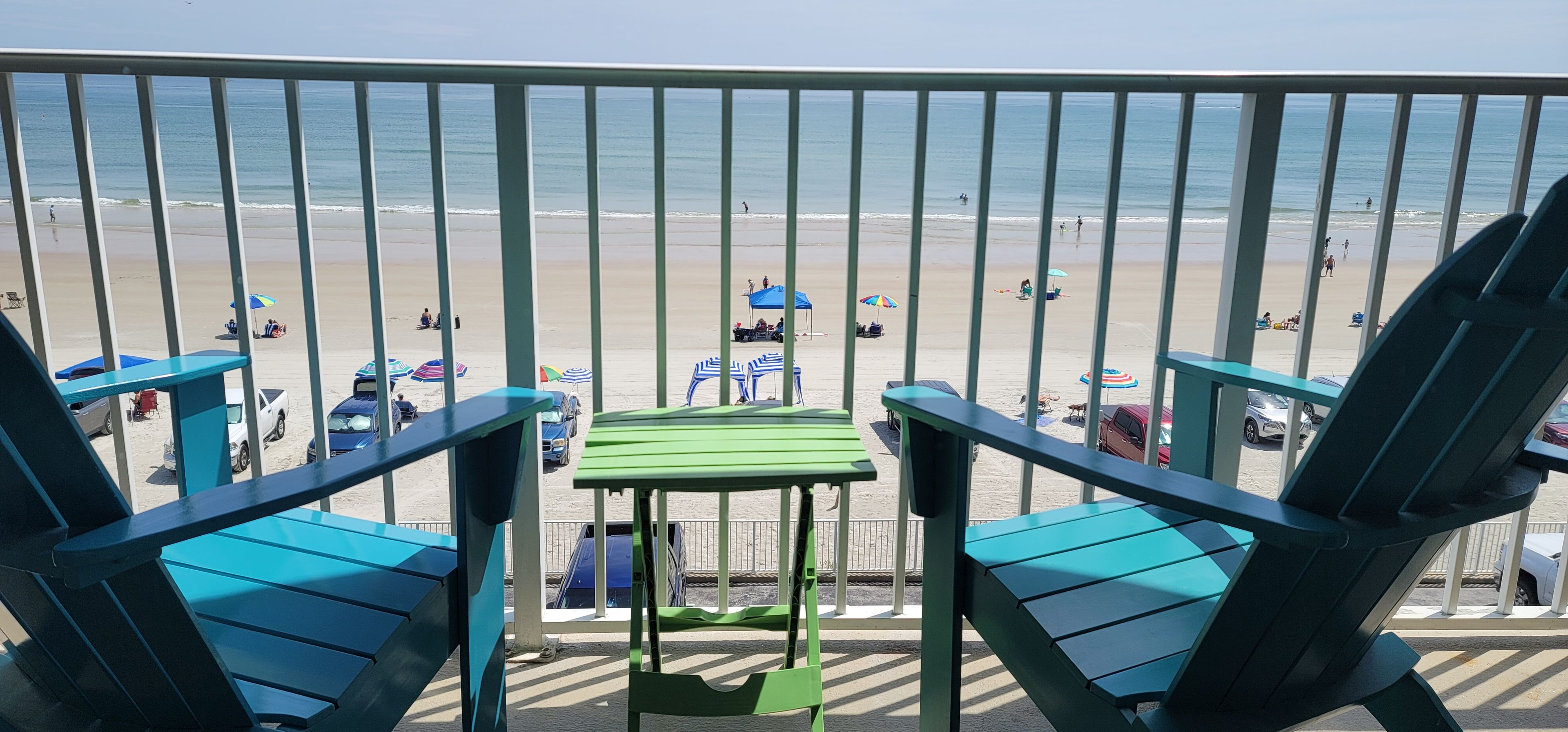 A balcony with two blue chairs and a small table, overlooking a beach with umbrellas and people.