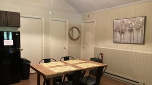 Uma sala de jantar com uma mesa de madeira, quatro cadeiras, uma geladeira preta e um quadro na parede.