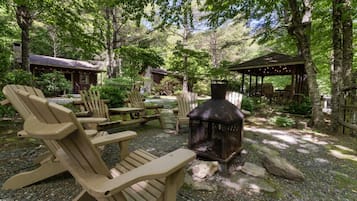Un espacio al aire libre rodeado de bosque, con sillas Adirondack, un hogar a leña y construcciones de madera.