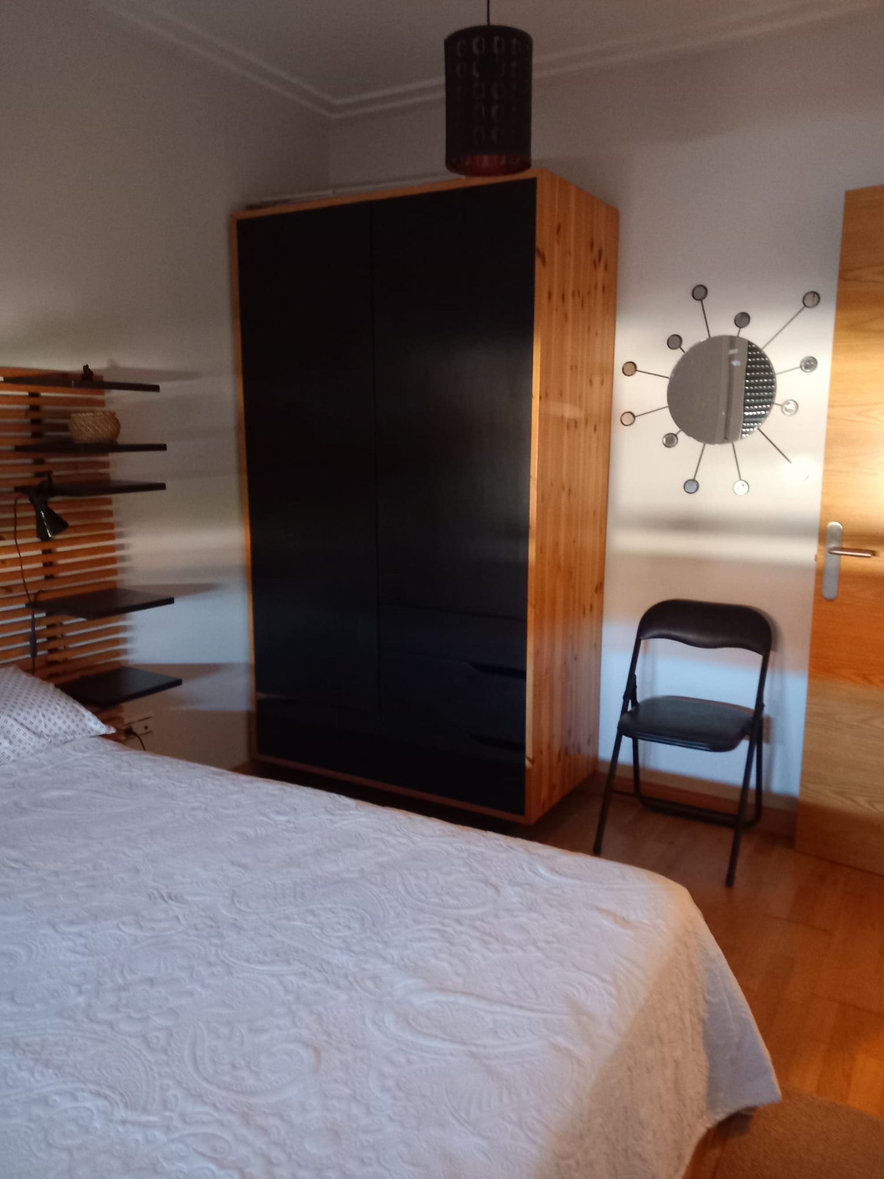 A bedroom with a bed, a wardrobe, a chair, and a wall-mounted light fixture.