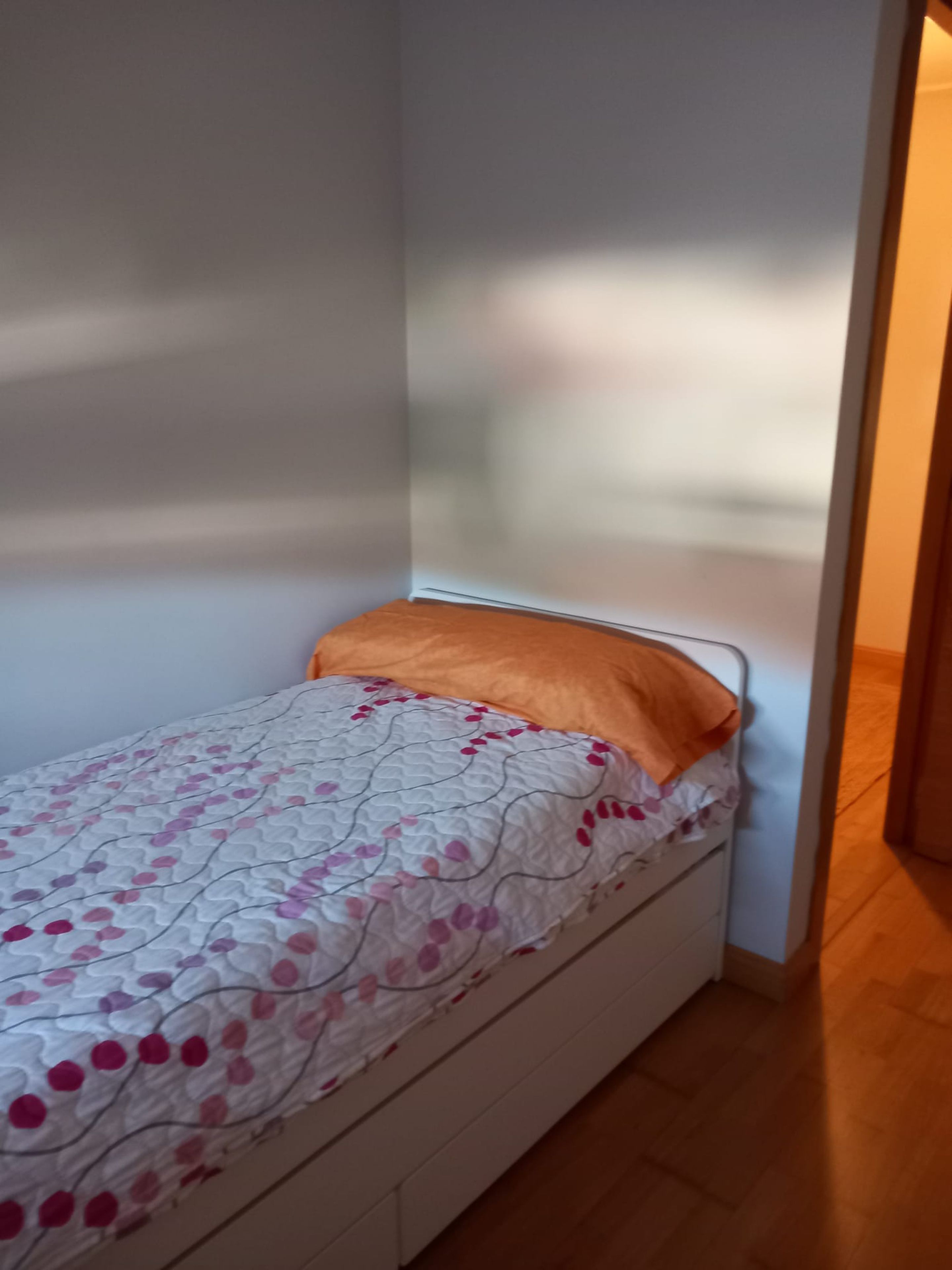 Una habitación pequeña con cama individual, pared blanca y suelo de madera.
