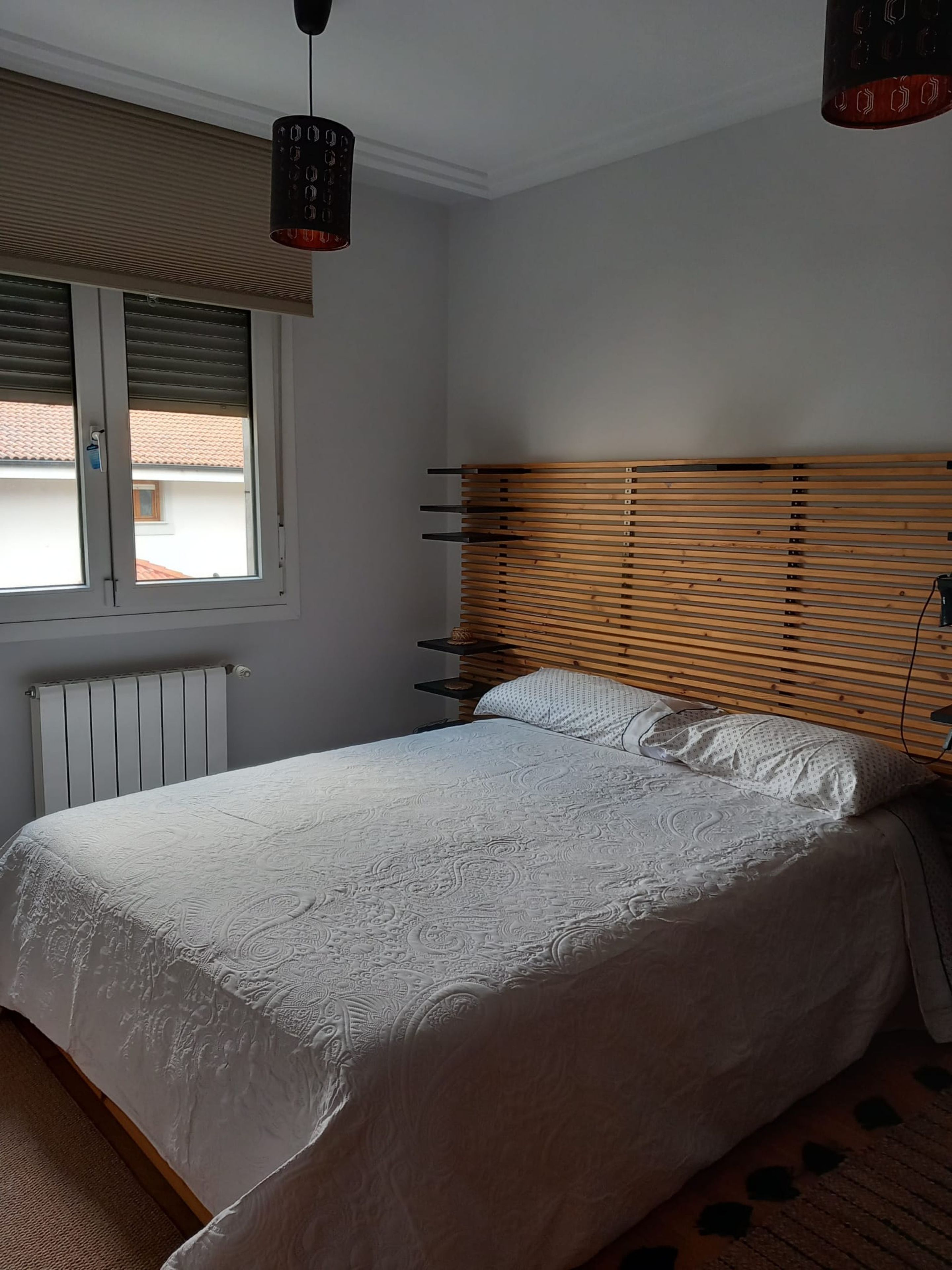 Un dormitorio con cama, ventana con persianas, radiador y cabecero de madera.