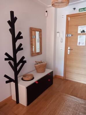 Un área de recepción con una puerta de madera, un espejo montado en la pared, una obra de arte abstracta negra, una cesta de mimbre y una pequeña consola blanca con cajones.