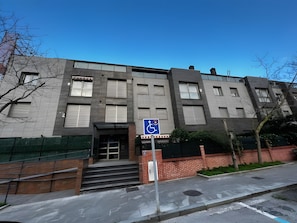 Un edificio de varios pisos con entrada accesible para sillas de ruedas, fachada de ladrillo y cielo azul despejado.