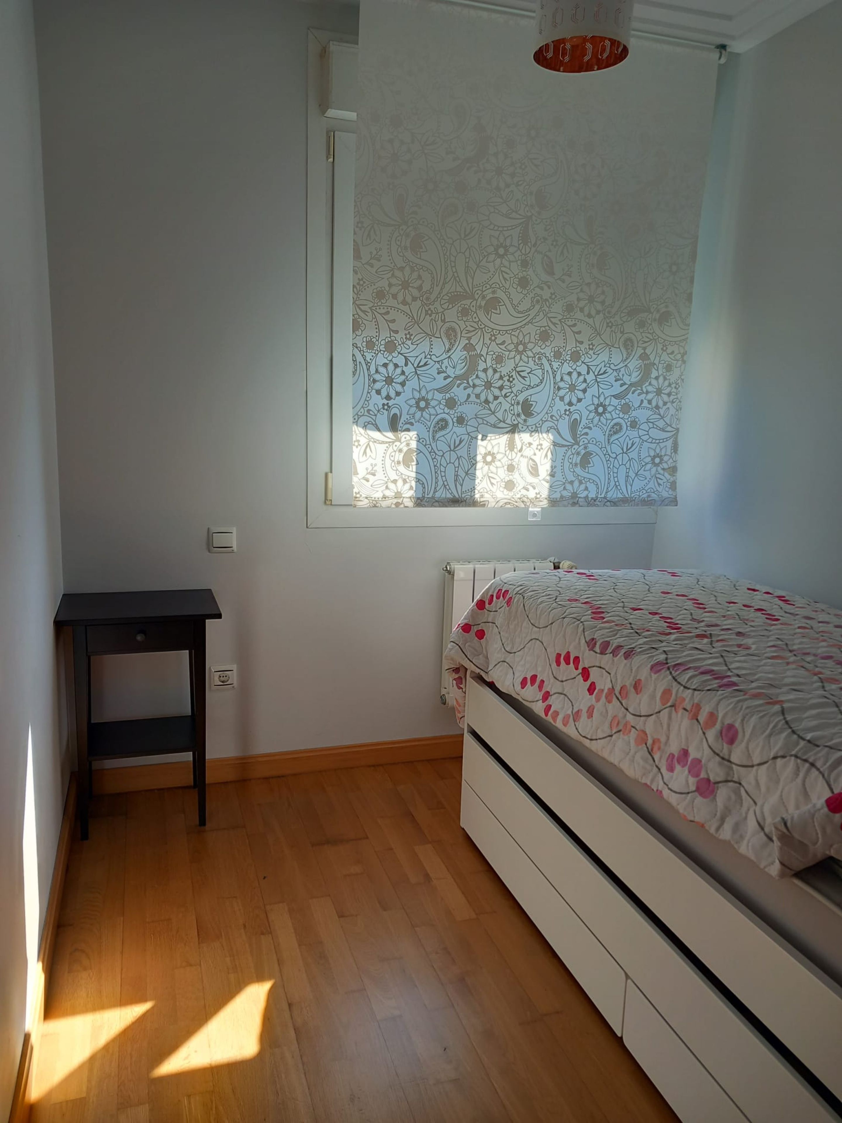 Un dormitorio con cama, una mesita, un radiador y una ventana con cortina estampada.