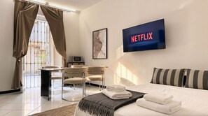 Ein Hotelzimmer mit einem Bett, einem Schreibtisch mit Stühlen, einem Fernseher, der 'NETFLIX' anzeigt, und einem Fenster mit Vorhängen.