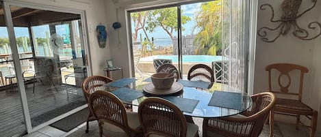 A dining area with a glass table and wicker chairs, overlooking a pool and greenery.