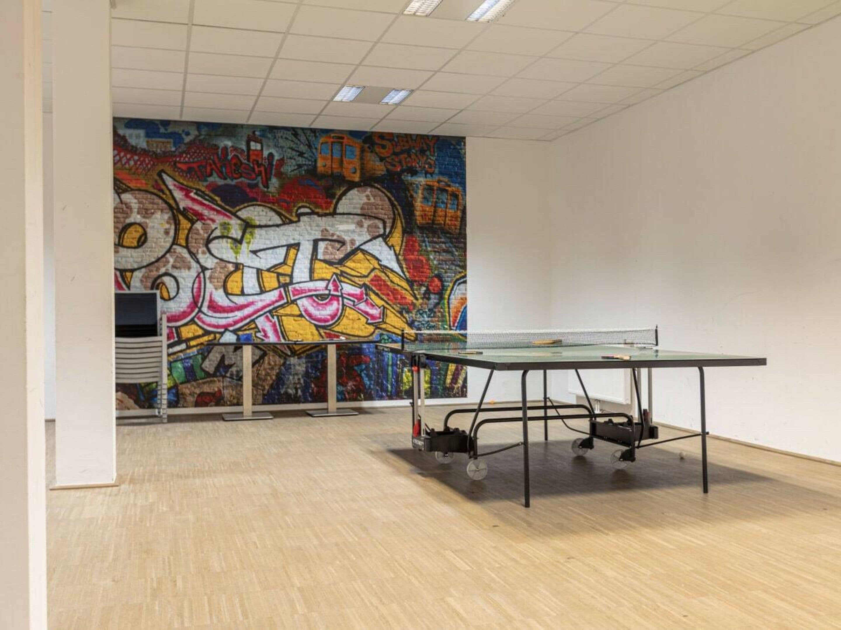 An indoor space with a ping pong table, a graffiti mural on the wall, and a whiteboard.