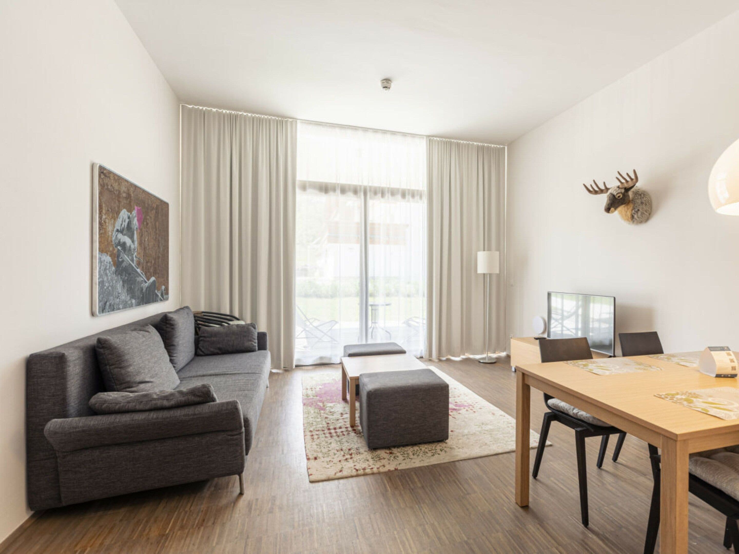 A modern living room with a grey sofa, a wooden dining table, and a large window with curtains.