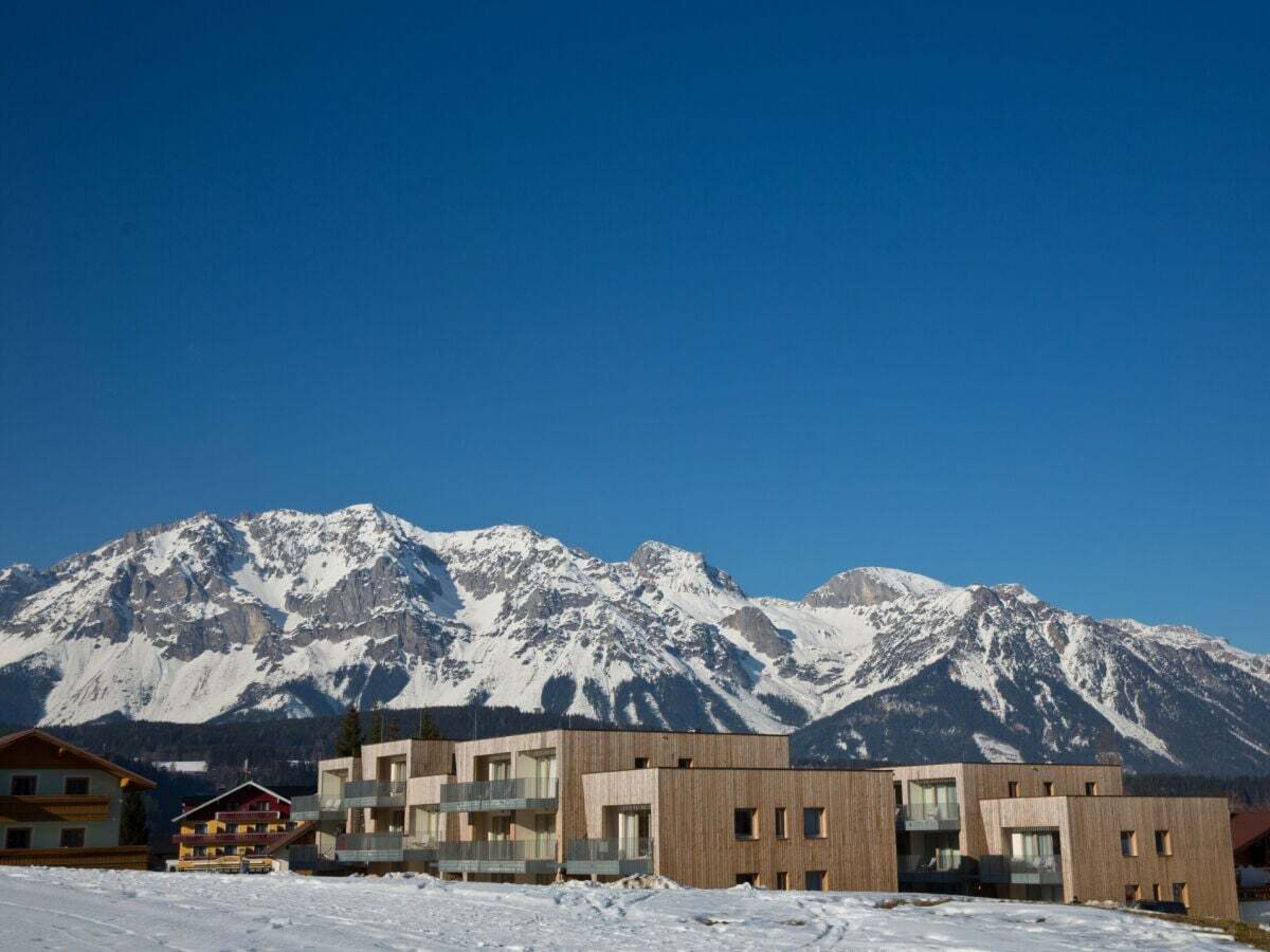 Ein modernes, mehrstöckiges Gebäude, vermutlich ein Hotel, mit Blick auf schneebedeckte Berge im Hintergrund.