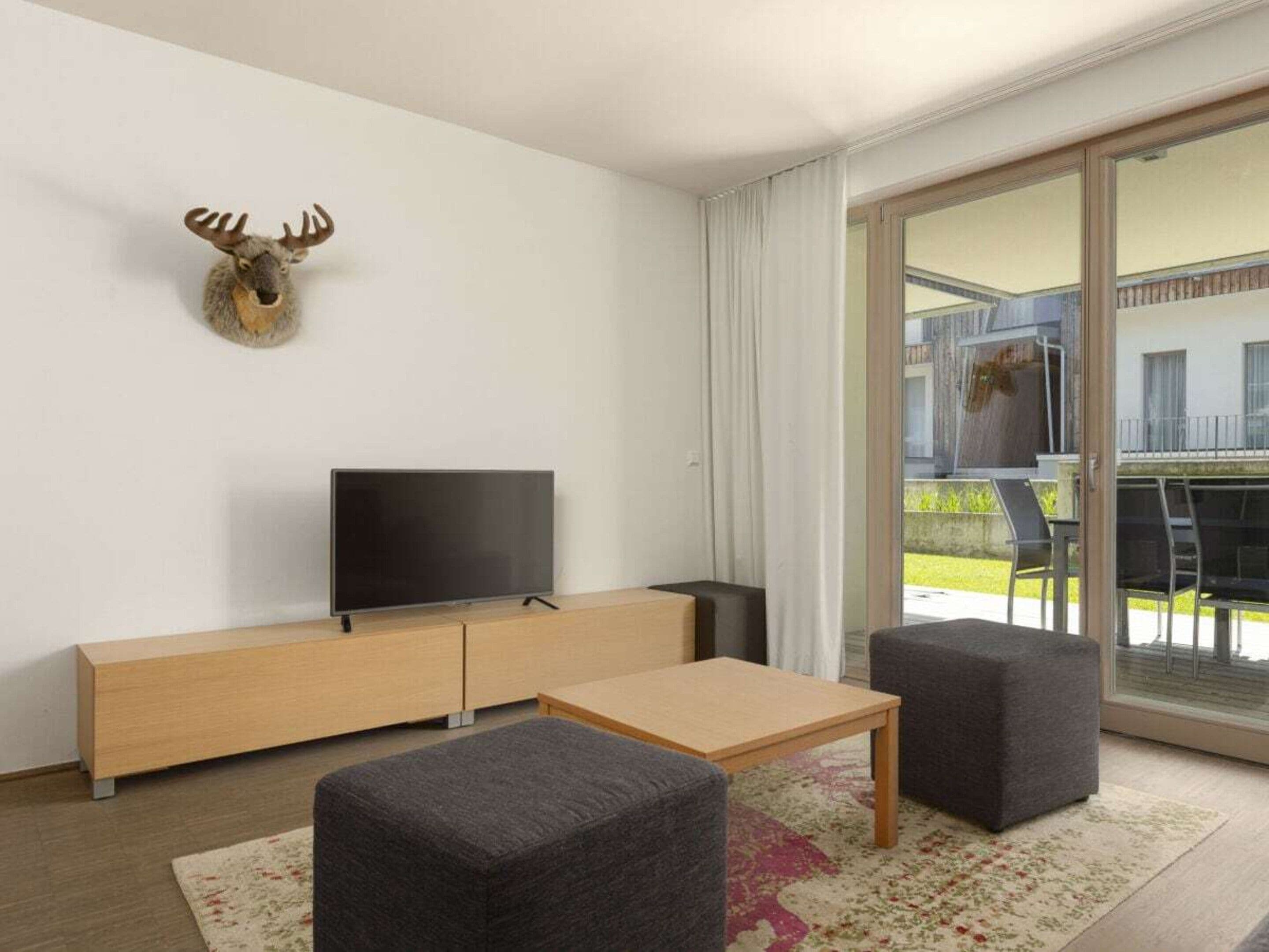 A modern living room with a flat-screen TV, a wooden cabinet, two grey ottomans, and a large sliding glass door leading to an outdoor area.