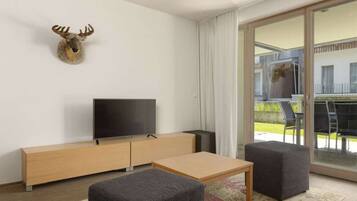 A modern living room with a flat-screen TV, a wooden cabinet, two grey ottomans, and a large sliding glass door leading to an outdoor area.