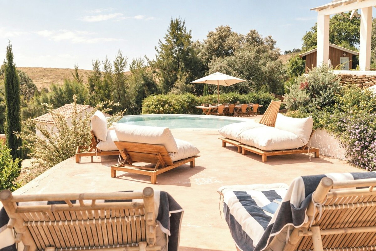 Zona junto a la piscina con sillones de descanso y una mesa, rodeada de vegetación y vistas al paisaje.