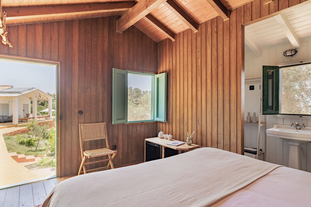 Un dormitorio con cama, una silla, una mesita con objetos y un baño con lavamanos.