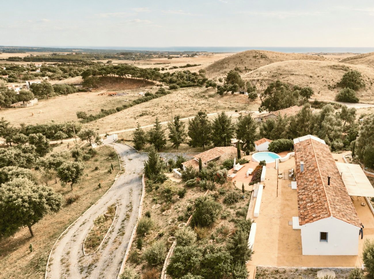 A rural landscape with a winding road, a white building with a tiled roof, and a swimming pool.