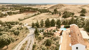 A rural landscape with a winding road, a white building with a tiled roof, and a swimming pool.