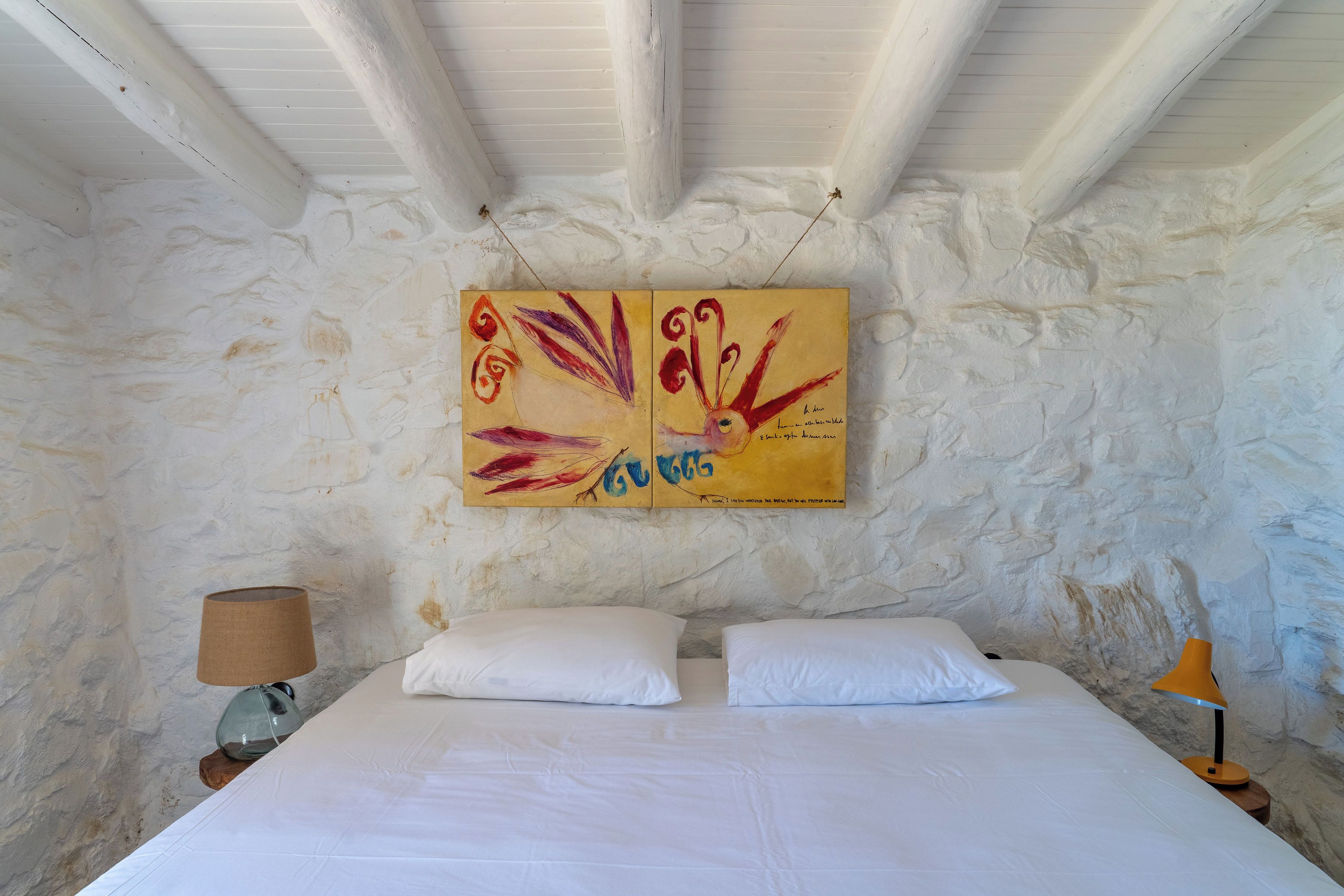 A bedroom with a bed, two pillows, a nightstand, a lamp, and a wall with a painting.