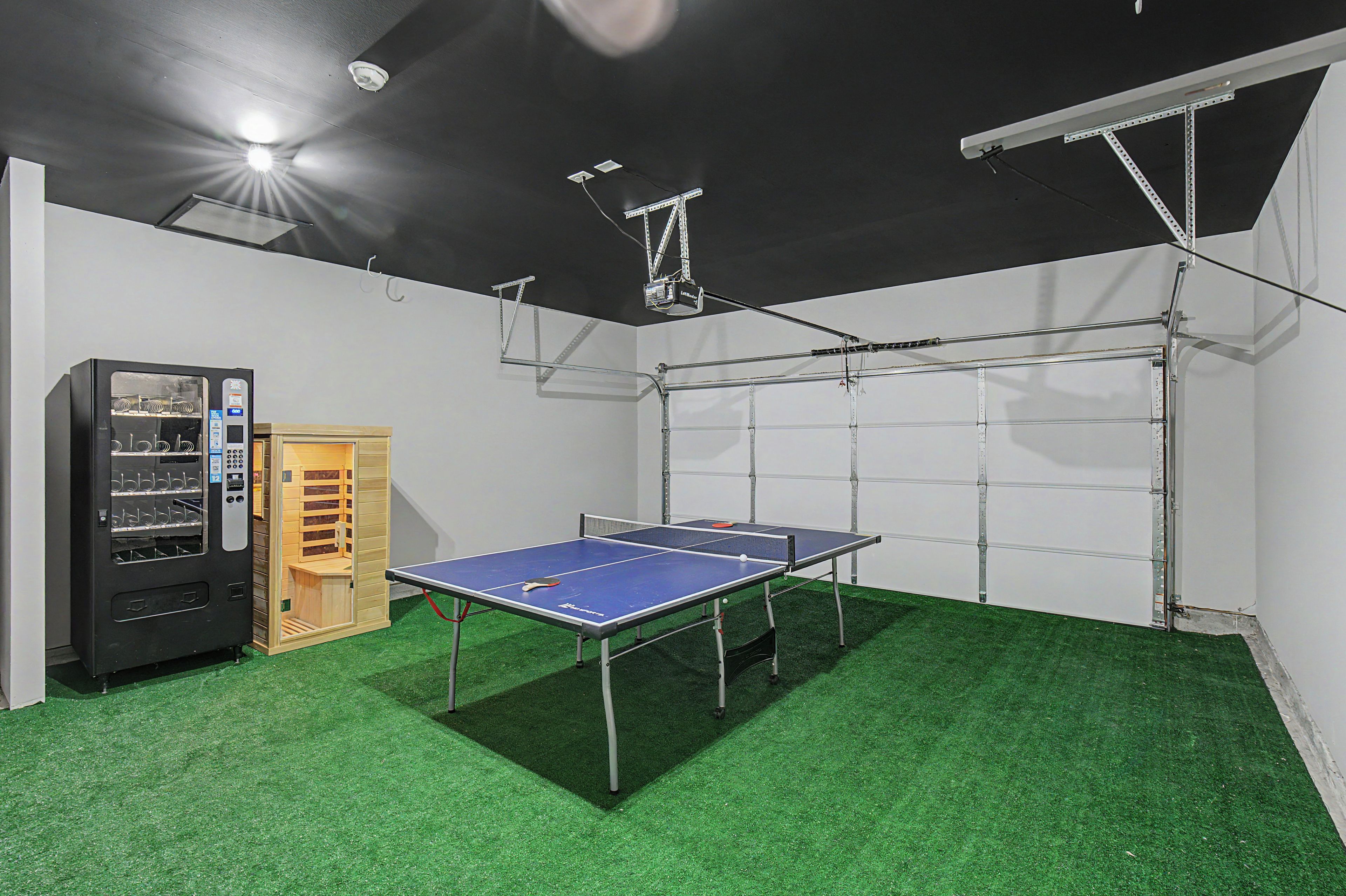 Une table de ping-pong avec un filet, un distributeur automatique et un meuble en bois, le tout dans une pièce dont le sol est recouvert de gazon synthétique.