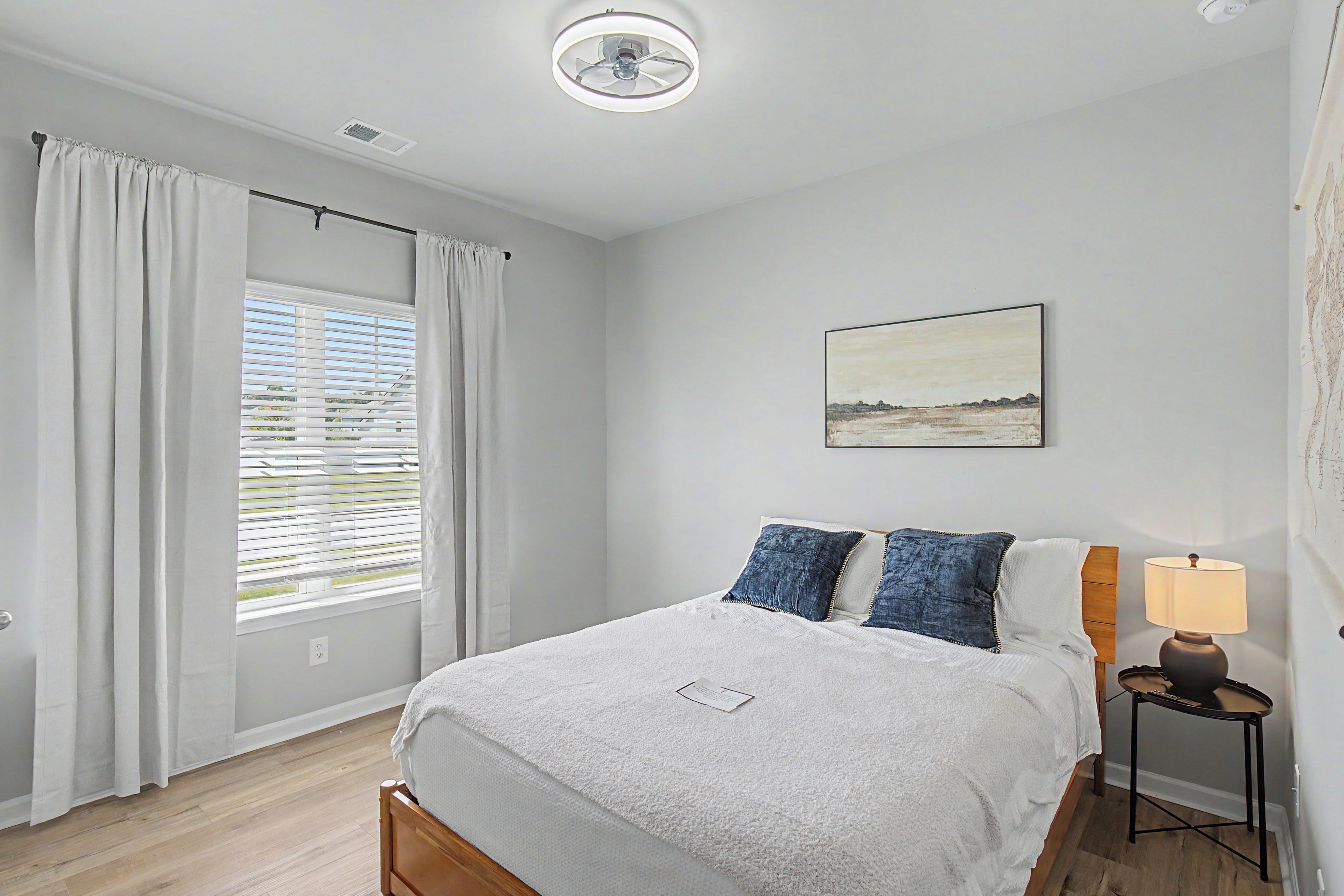 Une chambre à coucher avec un lit, une table de chevet avec une lampe, une fenêtre avec des stores et un tableau au mur.