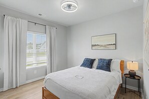 Ein Schlafzimmer mit einem Bett, einem Nachttisch mit Lampe, einem Fenster mit Jalousien und einem Bild an der Wand.