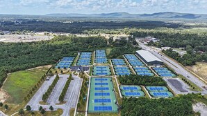 Une vue aérienne d’un complexe sportif comprenant plusieurs courts de tennis, des aires de stationnement et un grand bâtiment.