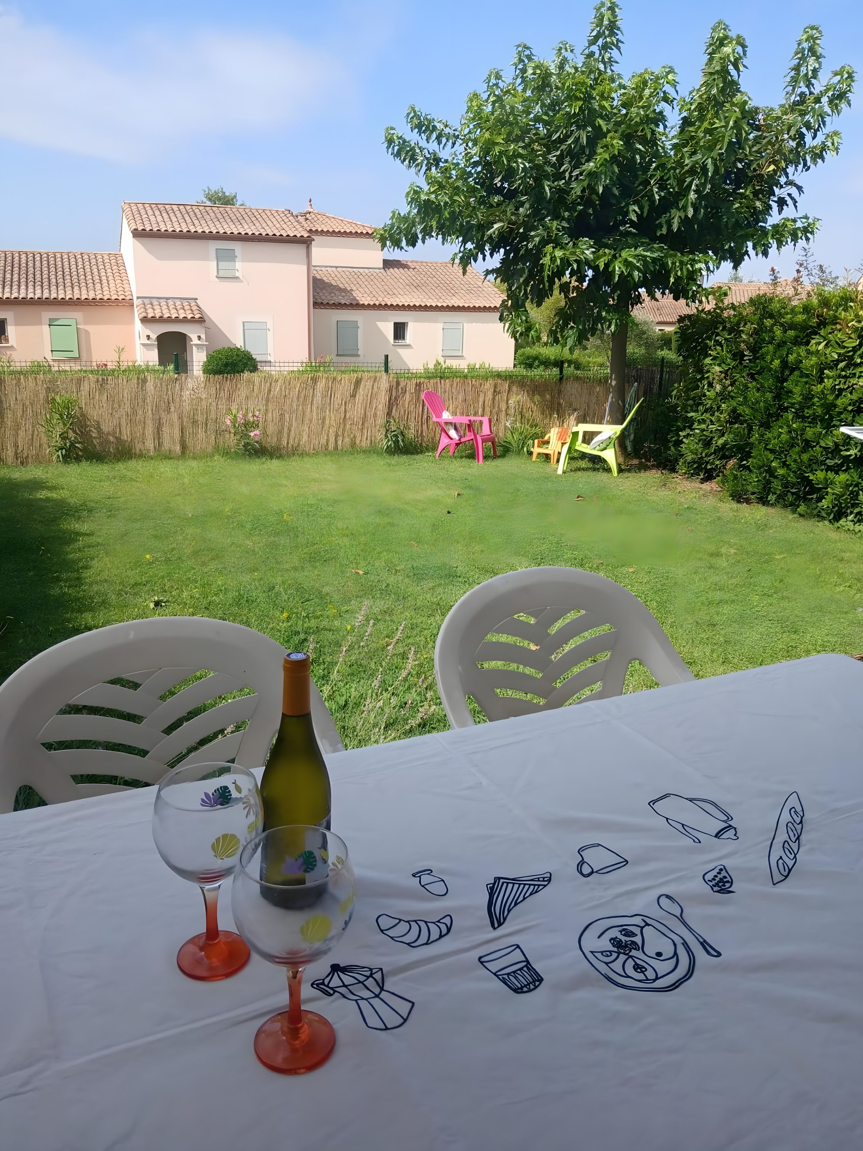 Een buitentafel gedekt, met een fles en glazen, omringd door stoelen, in een achtertuin met een gebouw op de achtergrond.