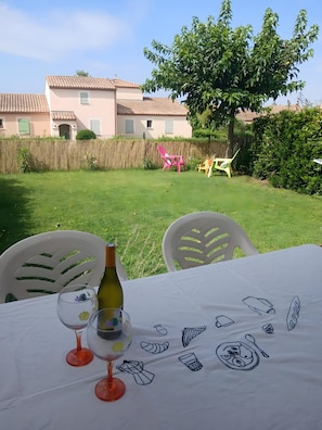 Une table dressée à l’extérieur, avec une bouteille et des verres, entourée de chaises, dans un jardin avec un bâtiment en arrière-plan.