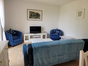 Un salon avec deux fauteuils bleus, un meuble TV blanc et un tableau accroché au mur.