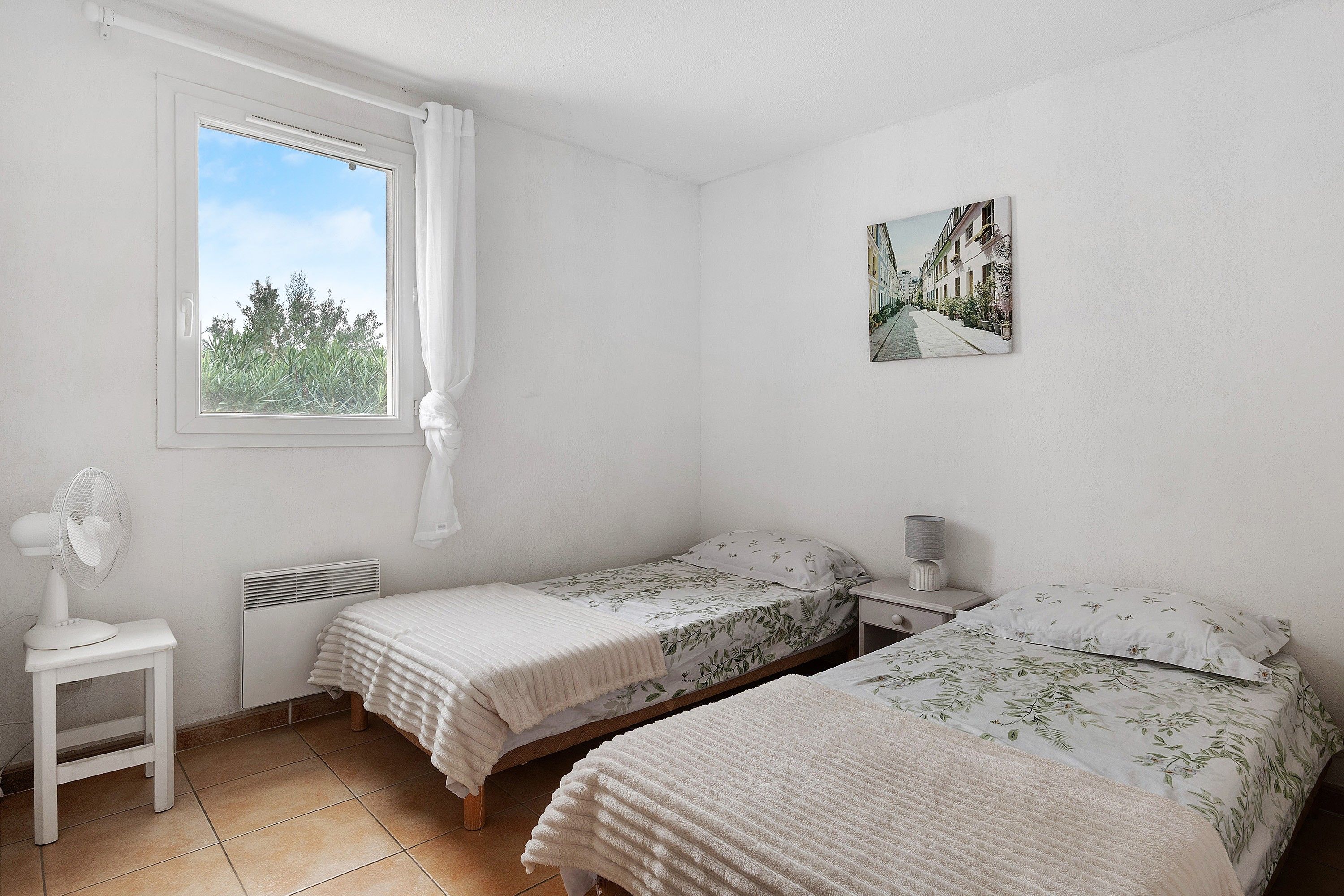 Une chambre avec deux lits, une fenêtre avec des rideaux, un radiateur, un ventilateur et un tableau accroché au mur.