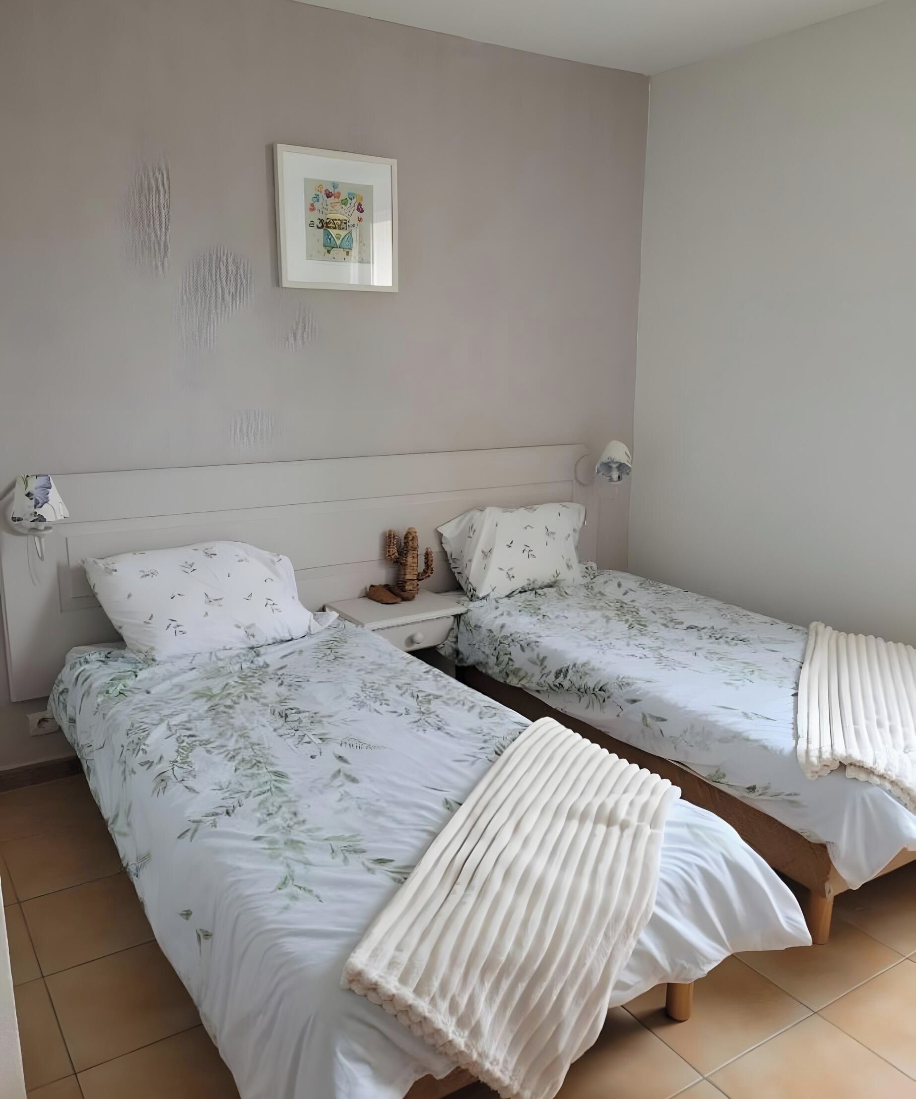 Ein Schlafzimmer mit zwei Einzelbetten, einem Nachttisch, einer Kakteen-Dekoration und einem gerahmten Bild an der Wand.