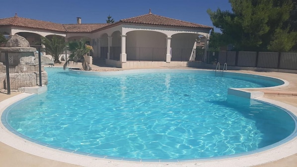 Une piscine à bord incurvé, entourée d’une terrasse carrelée et d’une structure de pavillon abrité.