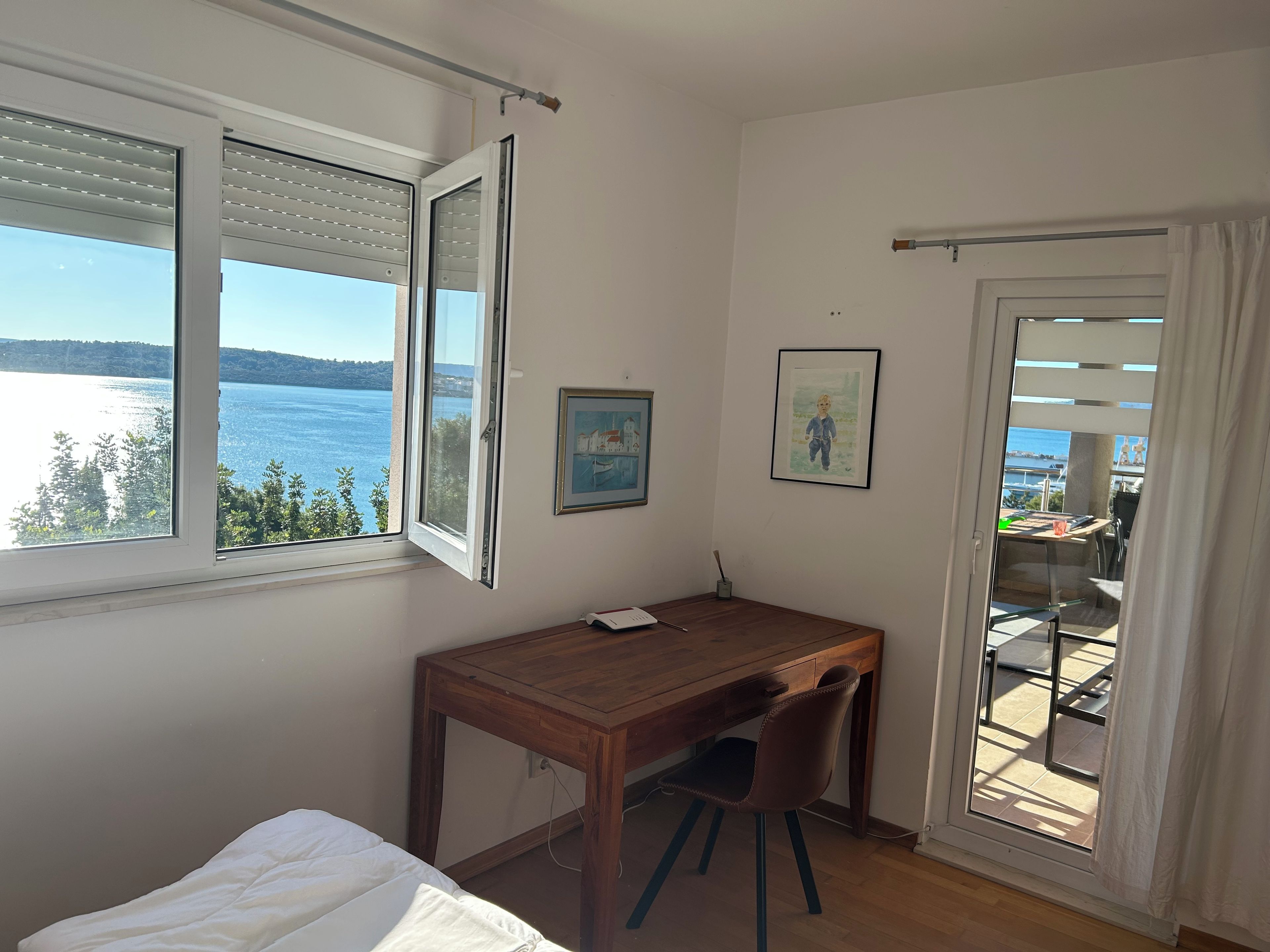 A room with a wooden desk, a chair, a bed, and a view of a body of water through a window.