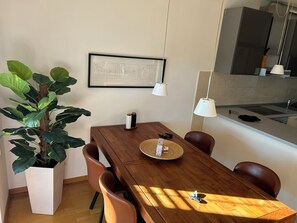 Ein Essbereich mit Holztisch, Stühlen, einem Blumentopf, einem gerahmten Bild und einer Küche im Hintergrund.