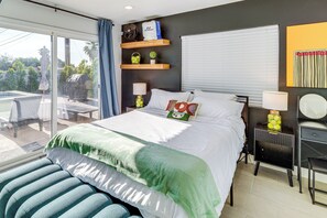A hotel room with a bed, a bedside table with a lamp, a window with blinds, a wall-mounted shelf with decorative items, and a view of the outdoors. - Remodeled Home w/ Mtn Views in Palm Springs! (Palm Springs)