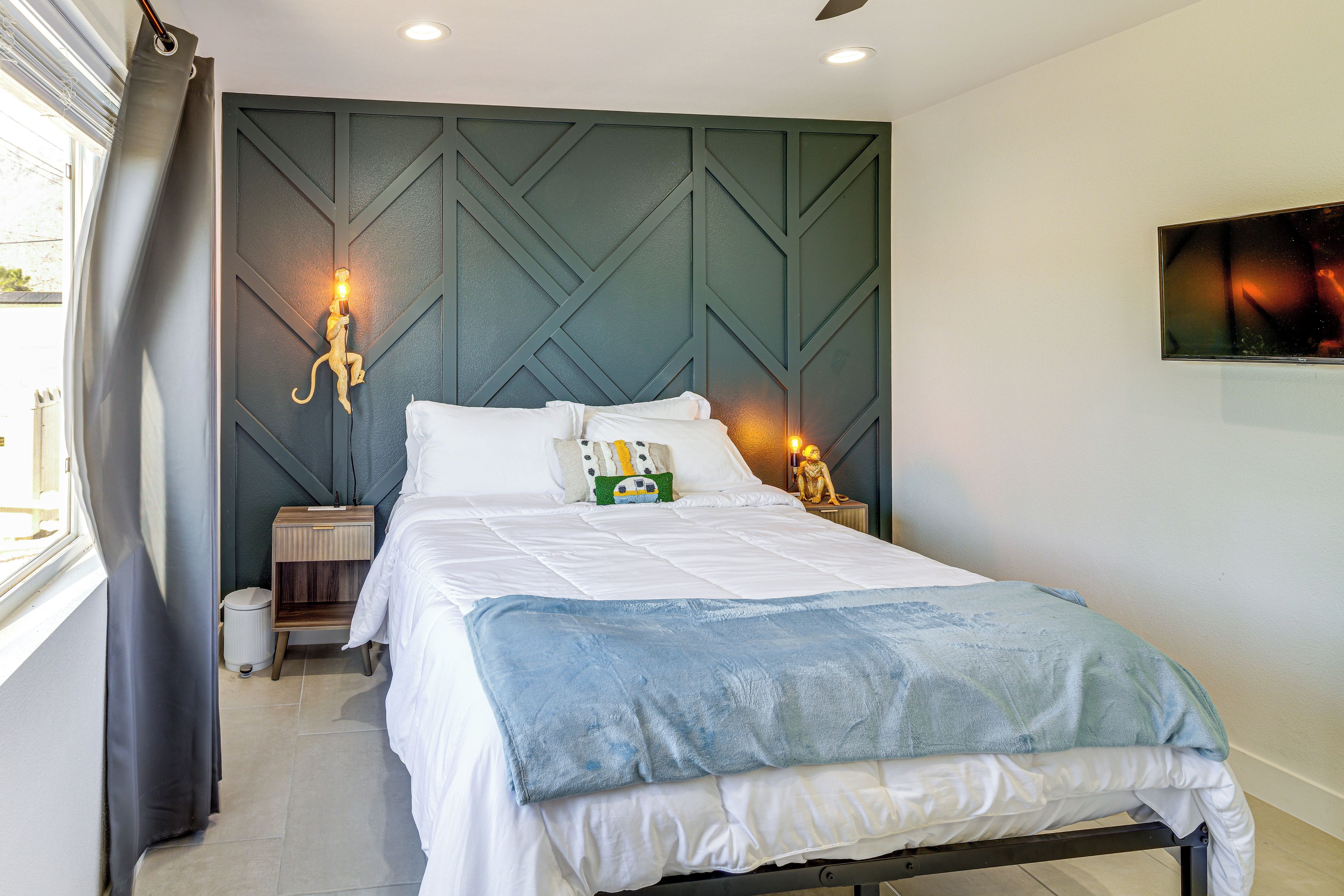 A neatly made bed with white linens and a blue throw blanket, a bedside table with a lamp, and a wall-mounted TV.
