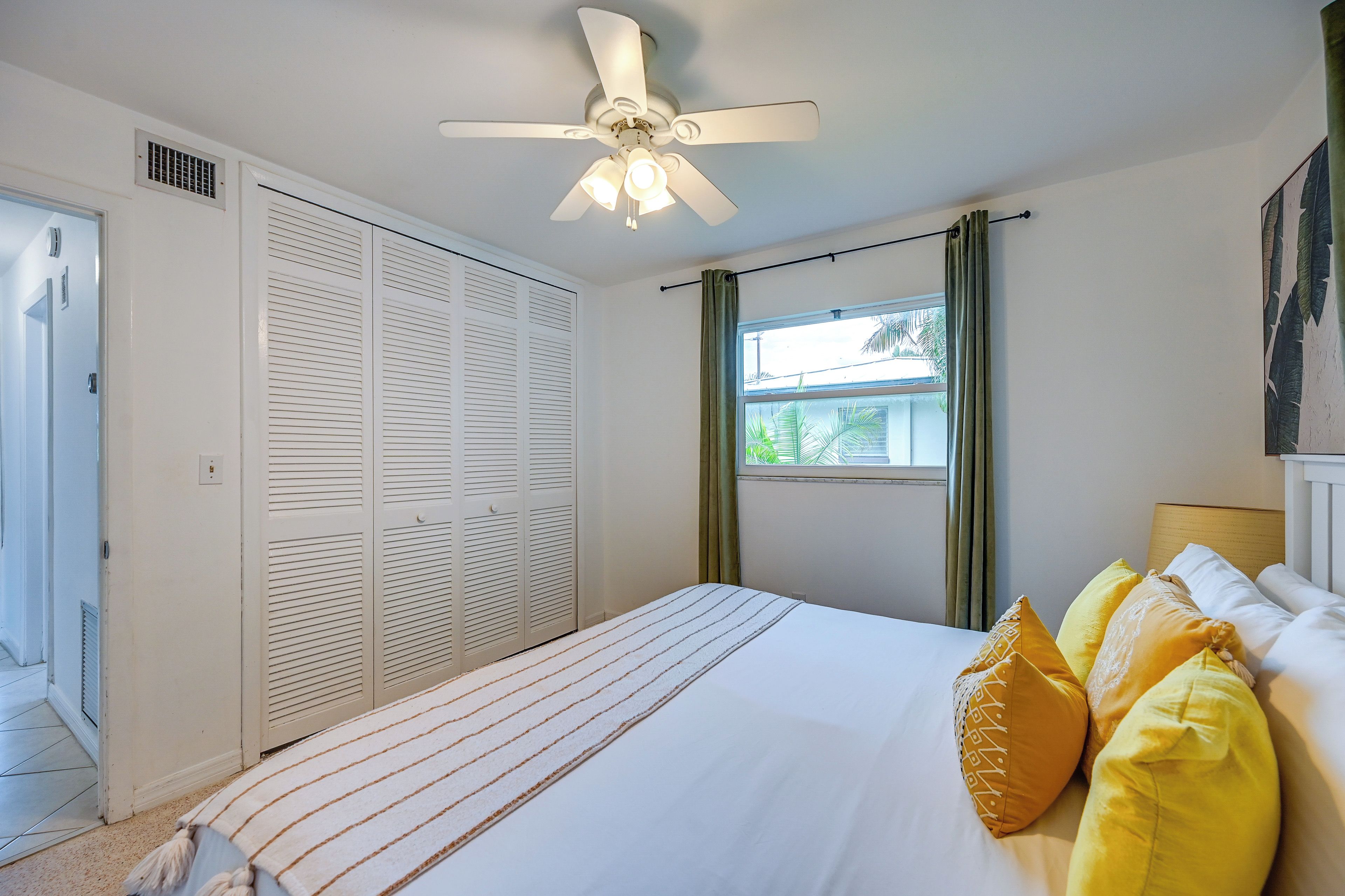 Ein Schlafzimmer mit einem Bett, einem Deckenventilator, einem Fenster mit Vorhängen und einer Tür mit Klappläden.