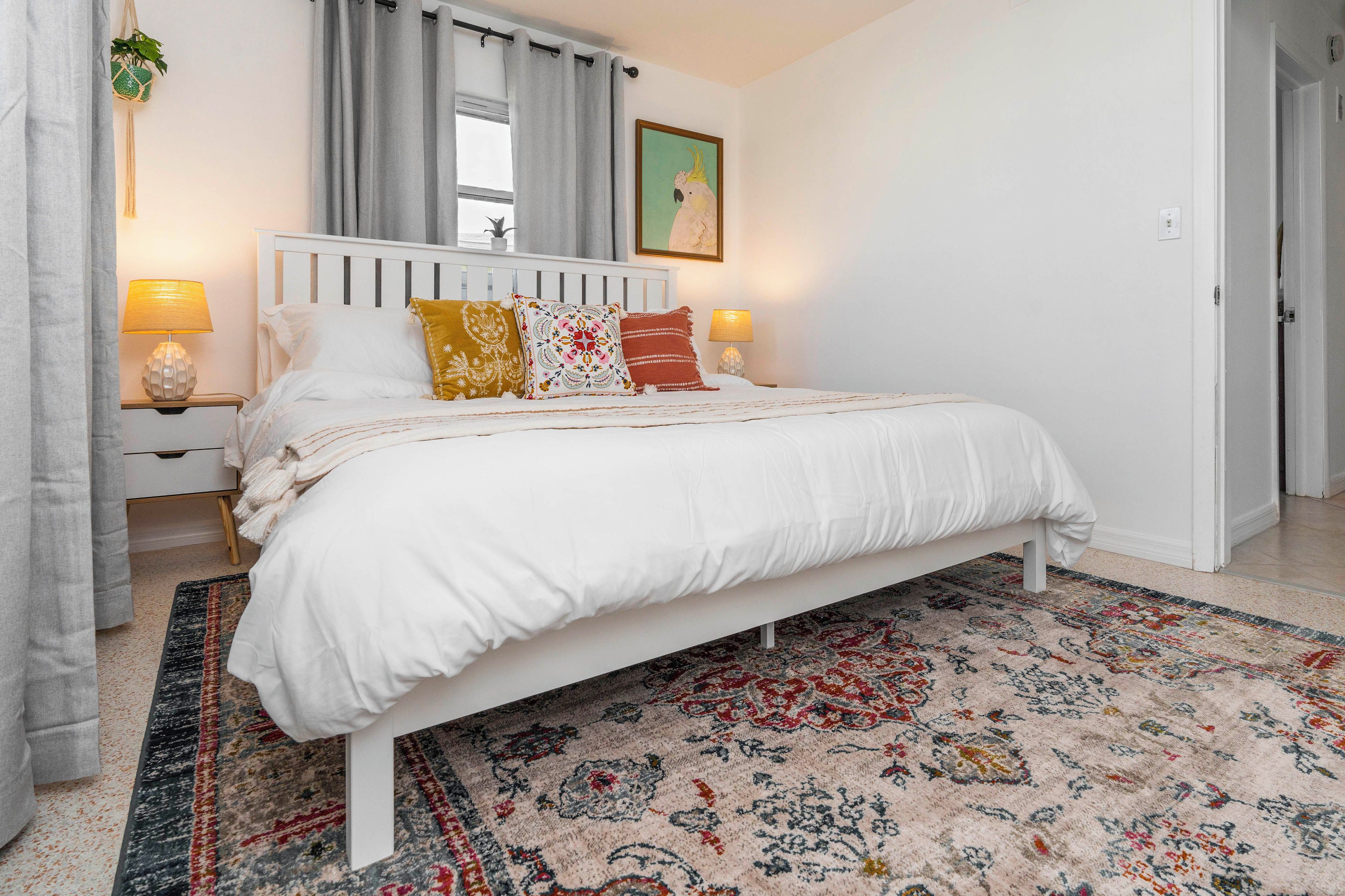A neatly arranged bedroom with a white bed, patterned pillows, a decorative rug, and a window with curtains.
