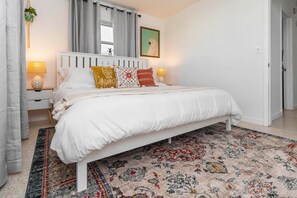 Ein ordentlich eingerichtetes Schlafzimmer mit einem weißen Bett, gemusterten Kissen, einem dekorativen Teppich und einem Fenster mit Vorhängen.